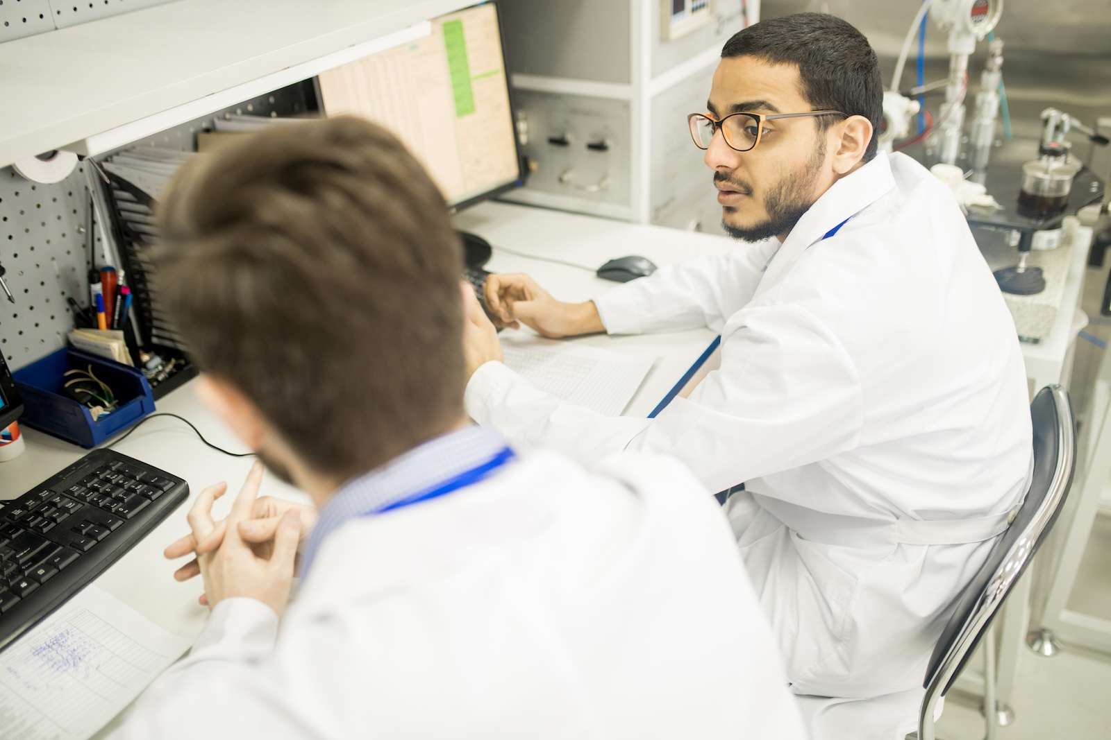 Multi-ethnic group of engineers wearing lab coats sitting at desk and discussing working process, interior of modern pressure transducers factory on background